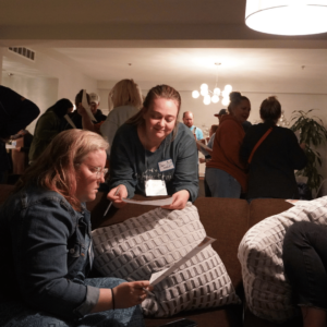 Two folks engaging together about a bingo card. One sitting on the couch and one standing leaning over the edge. People in the background standing and talking. 